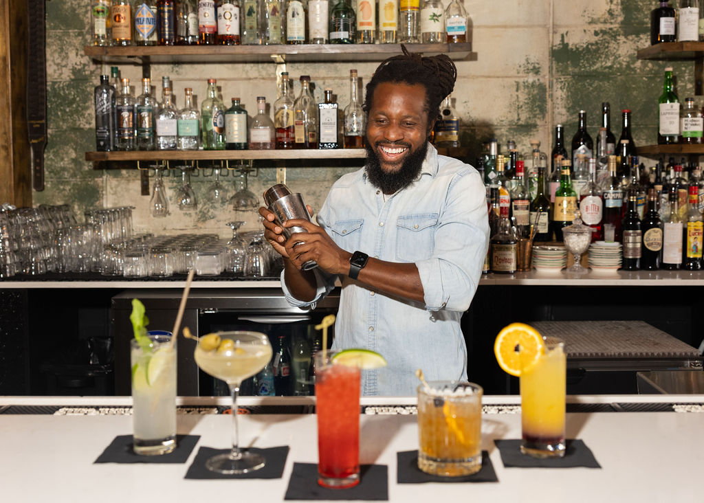 Daniel shaking a cocktail, laughing, with five drinks lined up in front